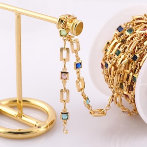 May include: A gold chain with square-shaped colourful gemstones. The chain is on a gold display stand and a spool of the chain is in the background.