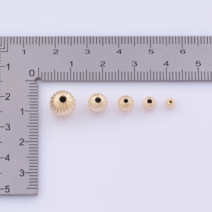 K&ouml;nnte beinhalten: Eine Reihe von sechs goldfarbenen, gerippten Perlen in verschiedenen Gr&ouml;&szlig;en. Die Perlen werden mit einem Lineal gemessen, wobei die gr&ouml;&szlig;te Perle etwa 0,6 cm Durchmesser hat. Jede Perle hat ein Loch in der Mitte.