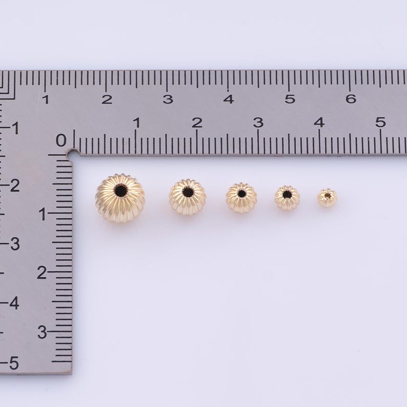 K&ouml;nnte beinhalten: Eine Reihe von sechs goldfarbenen, gerippten Perlen in verschiedenen Gr&ouml;&szlig;en. Die Perlen werden mit einem Lineal gemessen, wobei die gr&ouml;&szlig;te Perle etwa 0,6 cm Durchmesser hat. Jede Perle hat ein Loch in der Mitte.