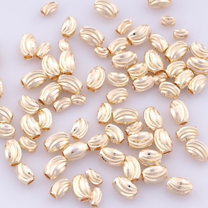 May include: A collection of gold-colored, barrel-shaped beads with horizontal ridged detailing. The beads vary in size and are scattered across a white surface, suitable for jewelry making or crafting projects.