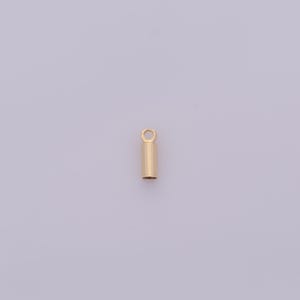 May include: A small, gold-colored cylindrical jewelry finding with a loop at the top. The pendant has a smooth, polished surface and is designed for attaching to a chain or other jewelry components. The background is a light purple color.