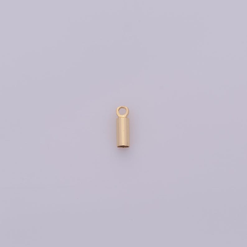 May include: A small, gold-colored cylindrical jewelry finding with a loop at the top. The pendant has a smooth, polished surface and is designed for attaching to a chain or other jewelry components. The background is a light purple color.