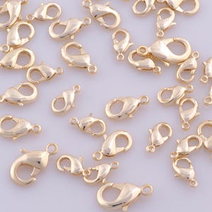 May include: A collection of gold-toned metal lobster claw clasps scattered across the frame. Each clasp features a spring-loaded mechanism and a small circular loop at one end for attachment, showcasing a smooth, polished, and reflective surface.