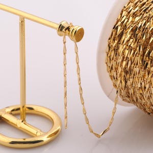 May include: A gold chain with a diamond-shaped pattern is displayed on a gold stand. The chain is wound around a spool.