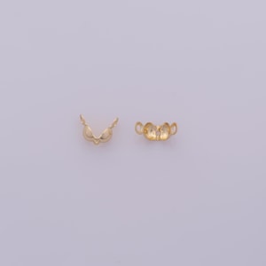 Peut inclure: Deux petites attaches de bijoux dor&eacute;es sont pr&eacute;sent&eacute;es sur un fond violet clair. L'une a une forme en V avec deux extensions incurv&eacute;es, l'autre a une forme arrondie avec trois boucles.