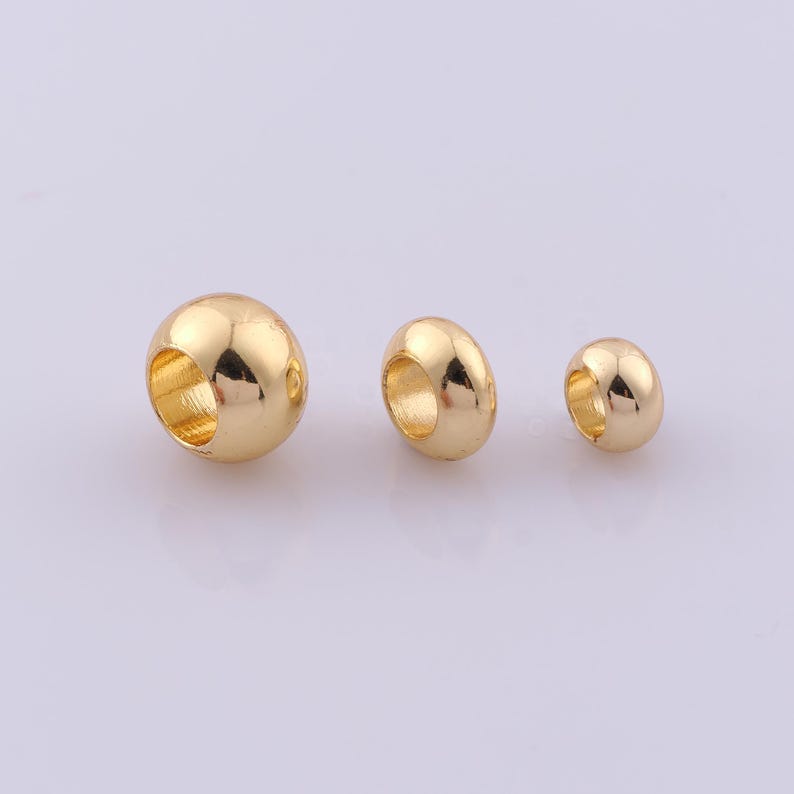 Peut inclure: Trois perles rondes et lisses en m&eacute;tal dor&eacute; de tailles diff&eacute;rentes dispos&eacute;es sur une ligne horizontale. Chaque perle pr&eacute;sente un trou central pour l'enfilage, avec une surface polie et r&eacute;fl&eacute;chissante qui capte la lumi&egrave;re, offrant un fini m&eacute;tallique.