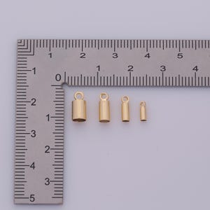 May include: Four gold-colored crimp end findings of varying sizes are arranged on a ruler. The findings are cylindrical with a loop at the top for attaching to jewelry. The ruler shows measurements in inches.