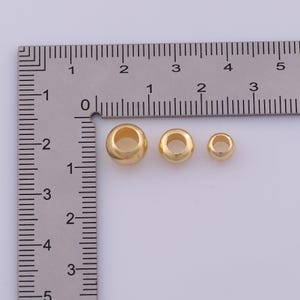 Peut inclure: Trois perles m&eacute;talliques lisses, rondes et de couleur dor&eacute;e, de tailles diff&eacute;rentes, dispos&eacute;es horizontalement &agrave; c&ocirc;t&eacute; d'une r&egrave;gle m&eacute;tallique en forme de L. La plus grande perle mesure environ 8 mm, la moyenne 5 mm et la plus petite 4 mm.