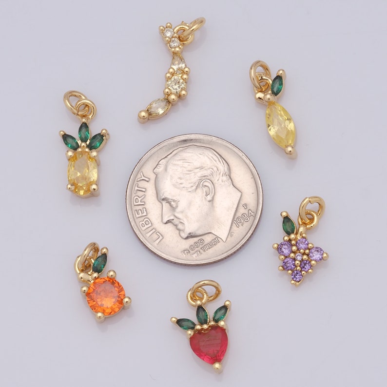 Op de afbeelding: Assortiment van goudkleurige bedels met verschillende vruchten, waaronder een ananas, een citroen, een appel, een aardbei en druiven. Elke bedel is versierd met gekleurde strass steentjes en groene emaille bladeren.
