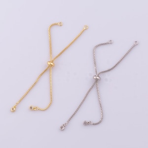 May include: Two adjustable bracelet chains, one in a gold tone and one in a silver tone. Each chain features a box link design, a sliding bead mechanism for size adjustment, and small circular end caps with a single embedded clear stone.