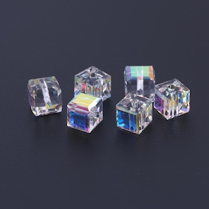 May include: Six clear, faceted crystal beads with iridescent color reflections. Each cube-shaped bead has a hole running through the center. The beads are arranged on a dark surface, showcasing their light-refracting properties.