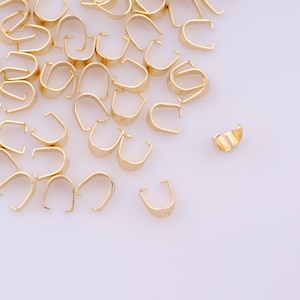May include: A selection of gold-coloured jewelry bails, used for connecting pendants to necklaces or bracelets. The bails feature a curved, U-shaped design with a small loop at the top for easy attachment. The bails are scattered on a white surface.