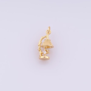 May include: A gold-colored charm in the shape of a small lamp. The lamp has a detailed lampshade, a decorative branch, and a star-shaped accent with a clear gemstone. The charm is designed for jewelry making.