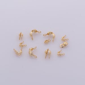 May include: A collection of small, gold-colored crimp end findings for jewelry making. These findings have a curved shape with a loop on one end and a split opening on the other, designed to secure the ends of jewelry cords or chains.