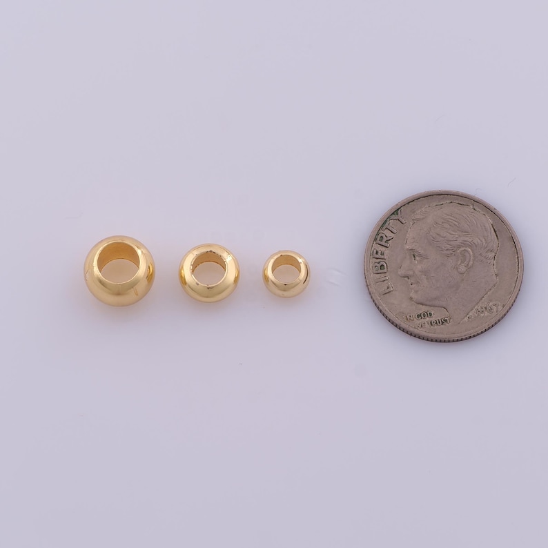 Peut inclure: Trois perles m&eacute;talliques lisses, rondes et de couleur dor&eacute;e, de tailles diff&eacute;rentes, dispos&eacute;es en rang&eacute;e horizontale &agrave; c&ocirc;t&eacute; d'une pi&egrave;ce de dix cents des &Eacute;tats-Unis, servant de r&eacute;f&eacute;rence d'&eacute;chelle pour la taille des perles.