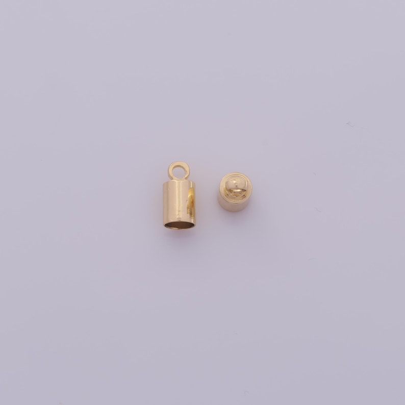 May include: Two gold-colored metal jewelry findings. One is a cylindrical crimp end with a loop, and the other is a small, rounded cap. These findings are used in jewelry making to secure the ends of cords or chains.