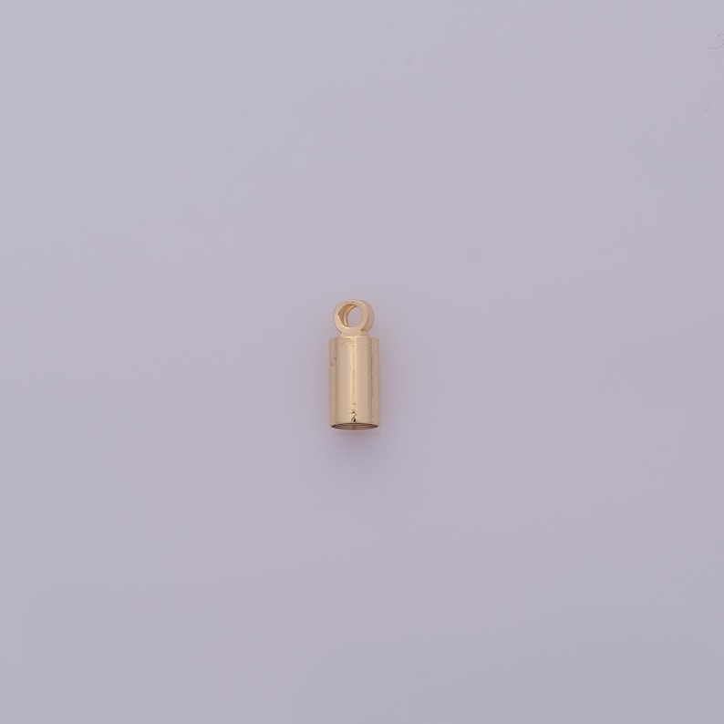 May include: A small, gold-colored cylindrical jewelry finding with a loop at the top. The finding is a simple, minimalist design, likely used for attaching pendants or charms to necklaces or bracelets.