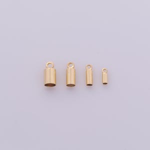 May include: Four gold-colored crimp end caps of varying sizes, each with a small loop at the top. The caps are cylindrical, with a smooth, polished finish. They are arranged horizontally against a plain, light background, suitable for jewelry making.