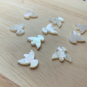 May include: A collection of ten white mother of pearl doves, each with a unique design, on a light wood surface.