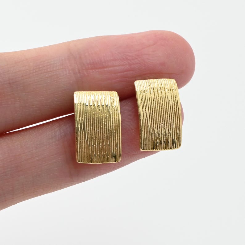 May include: A pair of gold-coloured rectangular stud earrings. Each earring has a textured, vertical lined pattern. The earrings are displayed against a light background.