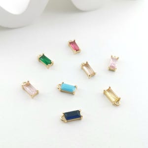 May include: Assortment of eight gold-toned charms with different colored rectangular glass stones. The charms have a loop at the top for attaching to jewelry.