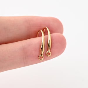 Op de afbeelding: Een paar goudkleurige oorhaken met een glad, gebogen druppelvormig ontwerp. Elke haak heeft een gepolijste metalen afwerking en een kleine cirkelvormige lus aan de onderkant voor het bevestigen van sieradenonderdelen.