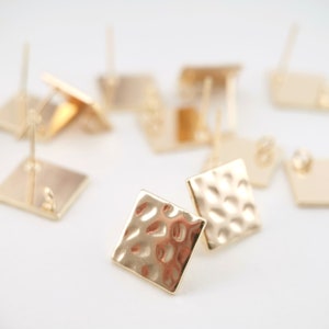 May include: A close-up of a group of gold-toned square earring studs with a hammered texture. The earrings are scattered on a white surface.