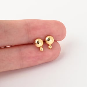 以下が含まれることがあります： ゴールドトーンのスタッドピアス一対。それぞれ上部に大きめの光沢のある球体ビーズがあり、そのすぐ下に小さめの同種の球体ビーズがぶら下がっている、反射する金属仕上げのシンプルな縦型デザインのピアスです。