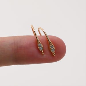 May include: A pair of gold-colored earrings with a curved hook design. Each earring features a teardrop-shaped embellishment with three small, clear gemstones. The earrings are displayed against a neutral background.