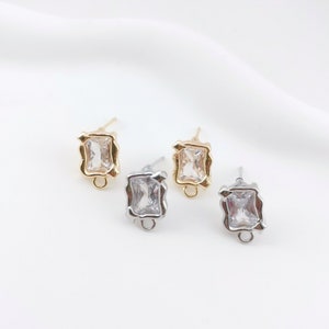 May include: Three pairs of earring findings with a clear square stone set in a gold or silver frame. The findings have a loop at the bottom for attaching a dangle earring.