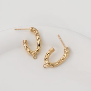 May include: A pair of gold-colored hoop earrings with a textured surface. The earrings have a post for piercing and a small loop at the bottom. The earrings are on a white surface.