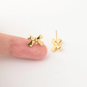 以下が含まれることがあります： 小さなリボンの形をしたゴールドトーンの金属製スタッドピアス一対。片方のピアスは指先に置かれ、もう片方はその横に立てられており、白い背景に対してポストとバタフライキャッチの留め具が見えています。