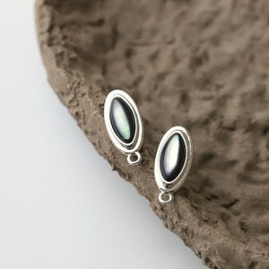 Pu&ograve; includere: Un paio di orecchini ovali color argento con un centro scuro e iridescente. Gli orecchini hanno un piccolo anello nella parte inferiore per il fissaggio. Lo sfondo &egrave; una superficie marrone strutturata.