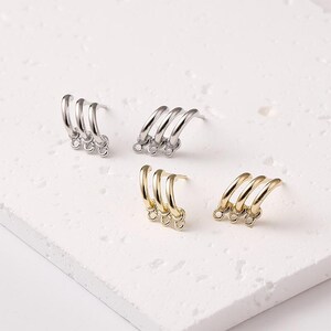 May include: Four pairs of earrings in silver and gold tones are displayed on a white surface. Each earring features three curved hoops connected at the bottom with small rings. The earrings are designed for pierced ears.