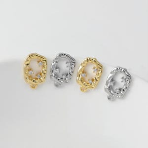 May include: Four small jewellery charms. Two are gold-toned, and two are silver-toned. Each charm has a rectangular shape with an irregular, textured border. Small, clear crystals are set within the charms, adding sparkle. Each charm has a small loop at the bottom.