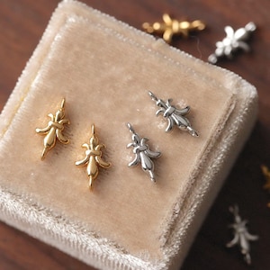 Può includere: Primo piano di diversi piccoli ciondoli decorativi in metallo. Due sono color oro e due color argento, tutti con un design a giglio. I ciondoli sono disposti su una superficie di velluto beige, con alcuni ciondoli in più visibili sullo sfondo.