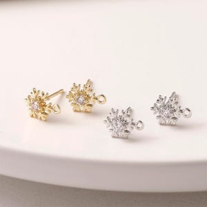以下が含まれることがあります： 4つの雪の結晶型のジュエリー。2つは金色、2つは銀色です。それぞれに中央に透明な宝石と、取り付け用の小さなループが付いています。イヤリングは白い表面にあります。