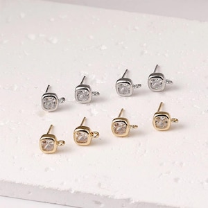 May include: A collection of square-shaped stud earring components. The earrings are available in silver and gold tones, each featuring a clear, square-cut gemstone. The components have a small loop for attaching additional elements.