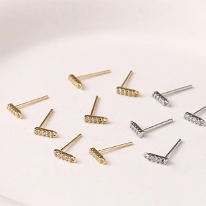 May include: A collection of gold and silver bar stud earrings. Each earring features a row of small, clear gemstones. The earrings are displayed on a white surface, showcasing their minimalist design and sparkle.