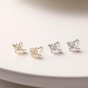 May include: Four starburst stud earrings. Two are gold-toned, and two are silver-toned. Each earring features a central, clear, round gemstone. The earrings have a small loop at the bottom for attaching to a chain or other jewelry component.