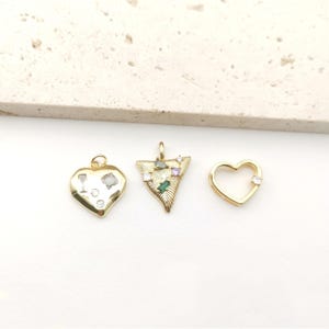 May include: Three gold-colored pendants. One is heart-shaped with clear and silver accents. Another is a triangle shape with green, purple, and clear stones. The third is a heart shape with a clear stone.