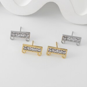 May include: Close-up of four stud earrings. Two are silver-toned, and two are gold-toned. Each earring features a rectangular bar design with five clear, square-cut stones. The earrings have a post and a small loop at each end.