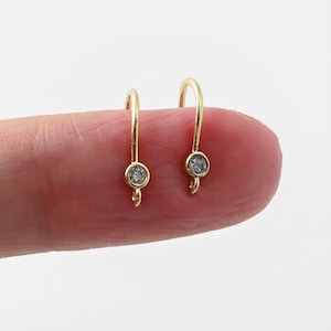 May include: Two gold-colored, small hoop earrings with a single, clear, round gemstone set in a bezel. The earrings have a small loop at the bottom for attaching charms or other decorative elements. The earrings are shown on a person's finger.