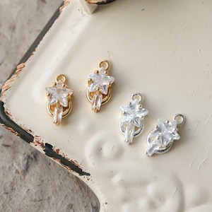 May include: Four star-shaped charms with clear crystals and gold or silver-colored metal. Each charm features a star-shaped crystal at the top, a rectangular crystal below, and a loop for attachment. The charms are displayed on a cream-colored surface.