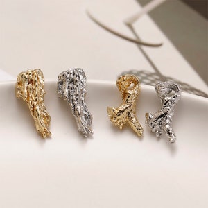 May include: Four textured metal charms in gold and silver. The charms have an organic, abstract shape, like a branch or coral. They are arranged on a white surface, with two gold and two silver charms.