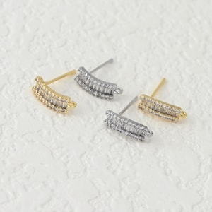May include: Four stud earrings with a curved bar design. Two are gold-toned, and two are silver-toned. Each earring is encrusted with small, clear crystals. The earrings are displayed on a textured, off-white surface.