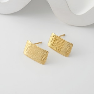 May include: A pair of gold-colored rectangular stud earrings. Each earring has a textured, ridged surface. The earrings are displayed on a white surface, with a soft, curved white shape in the background. Jewelry.