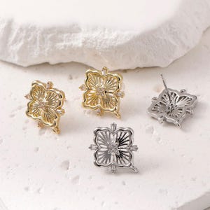 Pode incluir: Quatro brincos de botão em forma de flor em tons de ouro e prata. Os brincos dourados têm um acabamento brilhante com pequenos cristais transparentes. Os brincos prateados têm um acabamento fosco. Os brincos são exibidos em uma superfície branca texturizada.