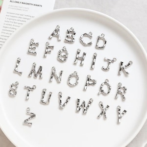 May include: A set of 26 silver metal alphabet letter charms. Each letter is individually crafted with a textured, hammered finish. The letters are arranged in alphabetical order on a white surface.