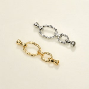 Pode incluir: Dois conectores de corrente de metal com um design oval texturizado e detalhes de cristal transparente. O conector à esquerda é dourado e o conector à direita é prateado.